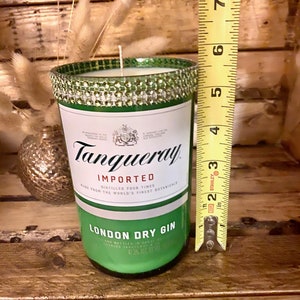 May include: A green glass candle jar with a silver and green glitter band around the top. The jar is labeled "Tanqueray Imported London Dry Gin" and has a measurement of 47.3% alc by vol. The jar is sitting on a wooden surface next to a measuring tape.