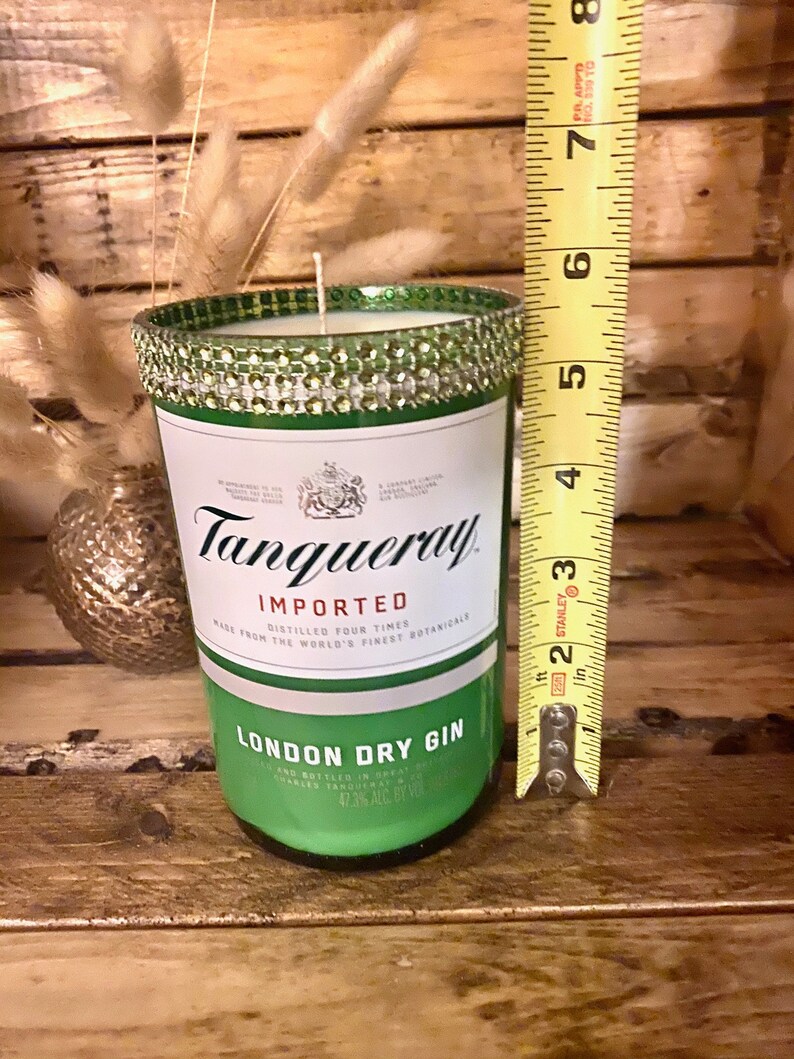 May include: A green glass candle jar with a silver and green glitter band around the top. The jar is labeled "Tanqueray Imported London Dry Gin" and has a measurement of 47.3% alc by vol. The jar is sitting on a wooden surface next to a measuring tape.