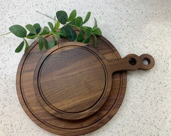 May include: A set of three round wooden serving boards, made of walnut wood. The boards are nested together, with the largest board having a handle. The boards are perfect for serving cheese, charcuterie, or other appetizers.