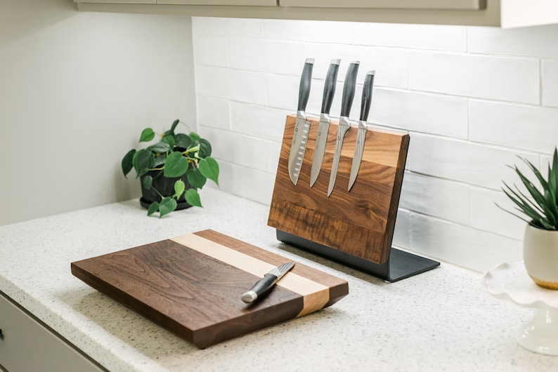 May include: A set of kitchen knives with black handles are magnetically attached to a wooden knife holder. A wooden chopping board with a light wood inlay is on the counter next to the knife holder.