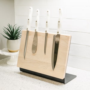 May include: A wooden magnetic knife holder with a black metal base, holding five knives with white handles. The holder is on a white countertop.