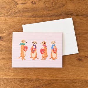 May include: A rectangular greeting card with a light pink background featuring horizontal lines and four illustrated meerkats. Each meerkat wears a colorful patterned scarf and holds a red heart, together spelling the word LOVE.