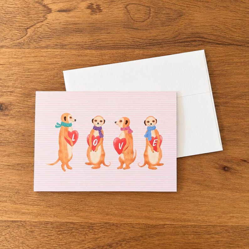 May include: A rectangular greeting card with a light pink background featuring horizontal lines and four illustrated meerkats. Each meerkat wears a colorful patterned scarf and holds a red heart, together spelling the word LOVE.