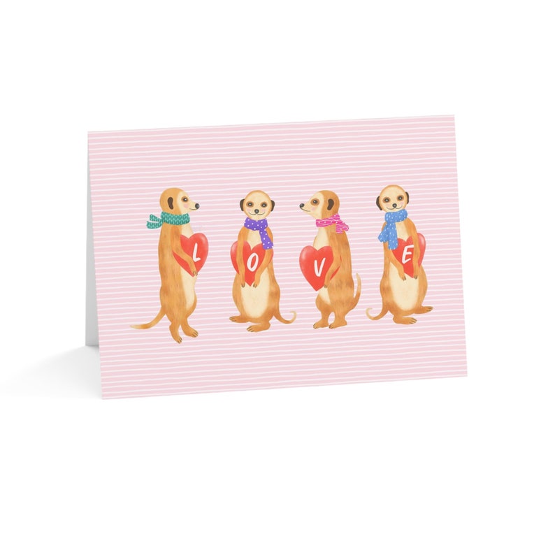 May include: A rectangular greeting card with a light pink background featuring thin white horizontal stripes. Four illustrated meerkats are standing in a row, each wearing a colorful patterned scarf and holding a red heart with a white letter spelling LOVE.