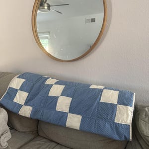 May include: A rectangular quilt featuring a checkerboard pattern of alternating solid off-white squares and blue squares with a small white polka dot print. The quilt is draped horizontally across the back of a gray sofa.