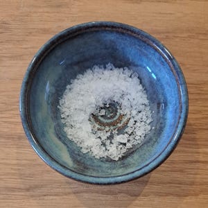May include: A small, round ceramic bowl with a variegated blue and brown glaze, filled with a portion of coarse white sea salt crystals. The interior of the bowl features a spiral pattern at the base, visible through the translucent salt.