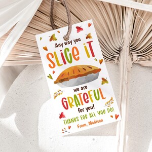 May include: A rectangular white gift tag with a brown leather string featuring a colorful illustration of a pie. The text reads: Any way you SLICE IT we are GRATEFUL for you! THANKS FOR ALL YOU DO! From, Madison. Small autumn leaves surround the text.