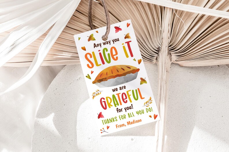May include: A rectangular white gift tag with a brown leather string featuring a colorful illustration of a pie. The text reads: Any way you SLICE IT we are GRATEFUL for you! THANKS FOR ALL YOU DO! From, Madison. Small autumn leaves surround the text.