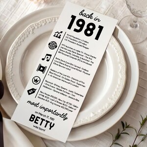 May include: A white card with black text, listing facts from 1981, rests on a white plate. The card reads "back in 1981" with facts about world population, MTV, movies, and music. The card also states "most importantly Betty was born!"