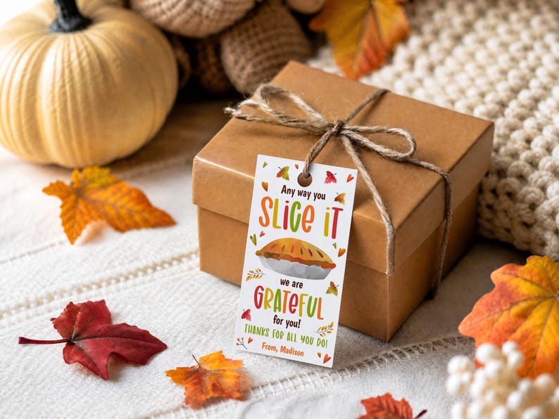 An editable Thanksgiving pie gift tag that says Any way you slice it we are grateful for you, featuring a watercolor pumpkin pie. The tag is tied with twine to a brown kraft gift box, sitting on a white knit blanket surrounded by autumn leaves and a white pumpkin.