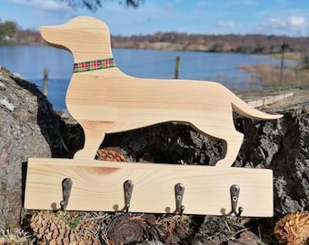 May include: Wooden dachshund key holder with five hooks. The dachshund is painted in a natural wood colour and has a red, green and yellow tartan collar. The key holder is sitting on a bed of pine cones.