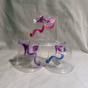 May include: Three clear plastic cups stacked on top of each other. Each cup contains a single, curled, fuzzy pipe cleaner. The top cup has a pink pipe cleaner, the middle cup has a purple pipe cleaner, and the bottom cup has a blue pipe cleaner.