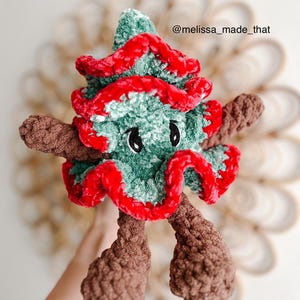 May include: A green and red crocheted Christmas tree plush toy with brown crocheted arms and legs. The toy has googly eyes and a red crocheted trim around the edges. The toy is held in a hand and the background is a blurry image of a woven basket.