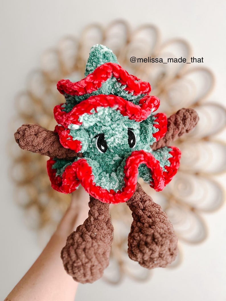 May include: A green and red crocheted Christmas tree plush toy with brown crocheted arms and legs. The toy has googly eyes and a red crocheted trim around the edges. The toy is held in a hand and the background is a blurry image of a woven basket.