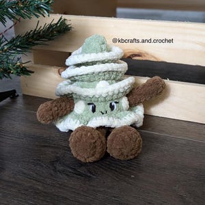 May include: A green crocheted Christmas tree plush toy with a white crocheted trim and brown crocheted legs. The toy has a smiling face with black eyes and a white mouth. The toy is sitting on a wooden surface. @kbcrafts.and.crochet