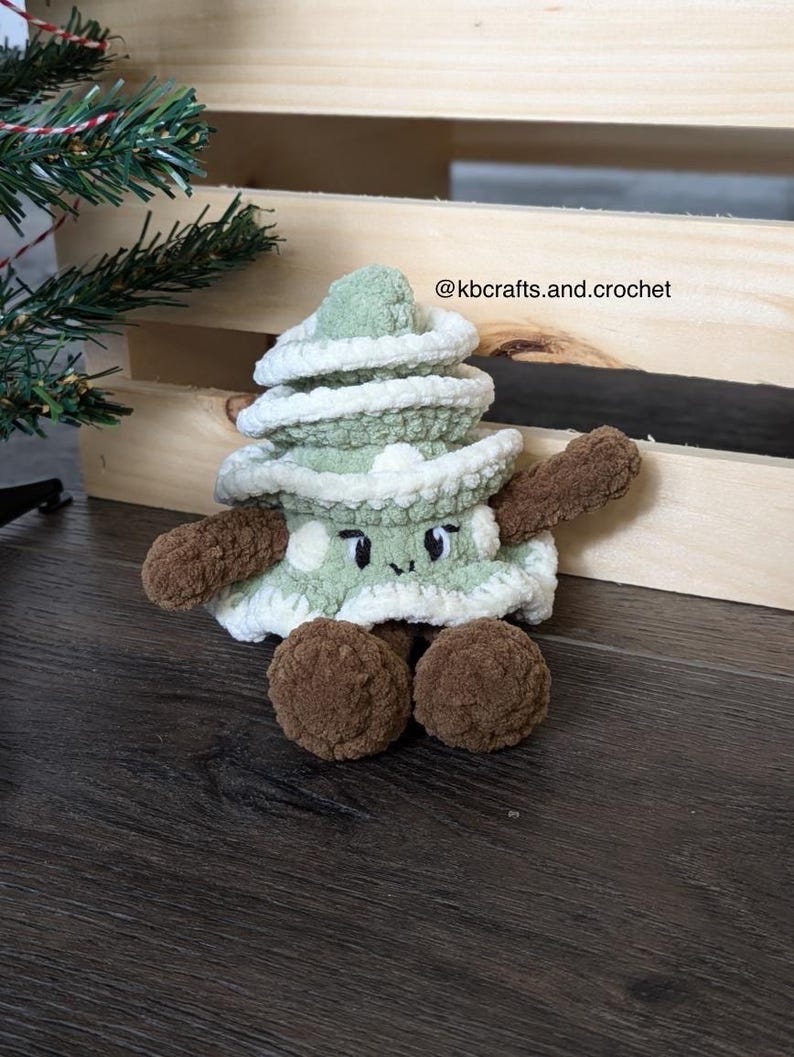 May include: A green crocheted Christmas tree plush toy with a white crocheted trim and brown crocheted legs. The toy has a smiling face with black eyes and a white mouth. The toy is sitting on a wooden surface. @kbcrafts.and.crochet