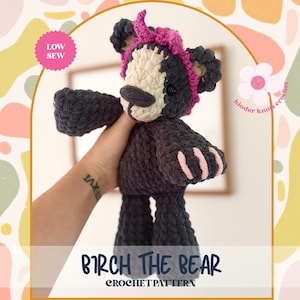 May include: A handmade crochet bear in dark gray with a cream-colored face and pink accents on the paws and headband. The bear has a dark gray nose and black eyes. The text "BIRCH THE BEAR CROCHET PATTERN" is displayed at the bottom.