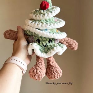 May include: A green crocheted Christmas tree toy with a red pom-pom on top. The tree has a white crocheted trim and a smiling face. The toy is being held by a hand wearing a pink smartwatch. The image includes the text "@smoky_mountain_lily".