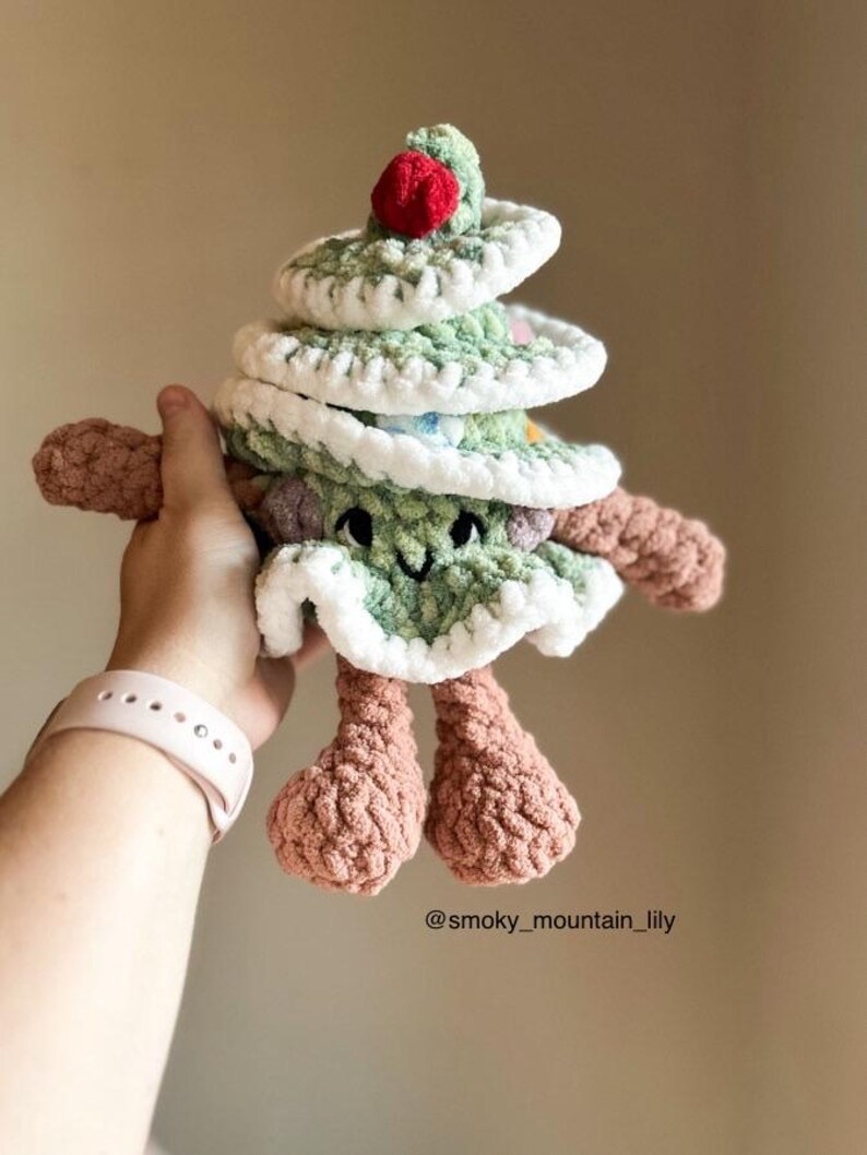 May include: A green crocheted Christmas tree toy with a red pom-pom on top. The tree has a white crocheted trim and a smiling face. The toy is being held by a hand wearing a pink smartwatch. The image includes the text "@smoky_mountain_lily".