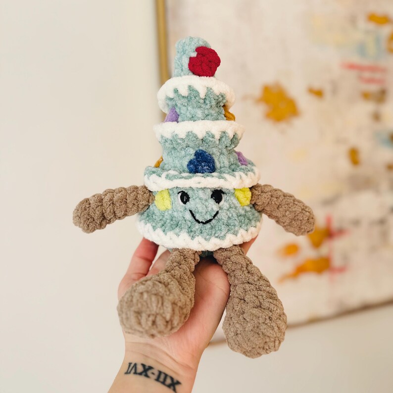 May include: A crocheted plush toy shaped like a Christmas tree with a smiling face. The tree is decorated with colorful yarn and a red pom-pom on top.