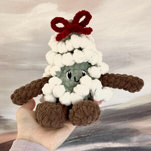 May include: A green crocheted Christmas tree plush toy with white pom-poms and a red bow on top. The toy has a smiling face and brown crocheted arms and legs.