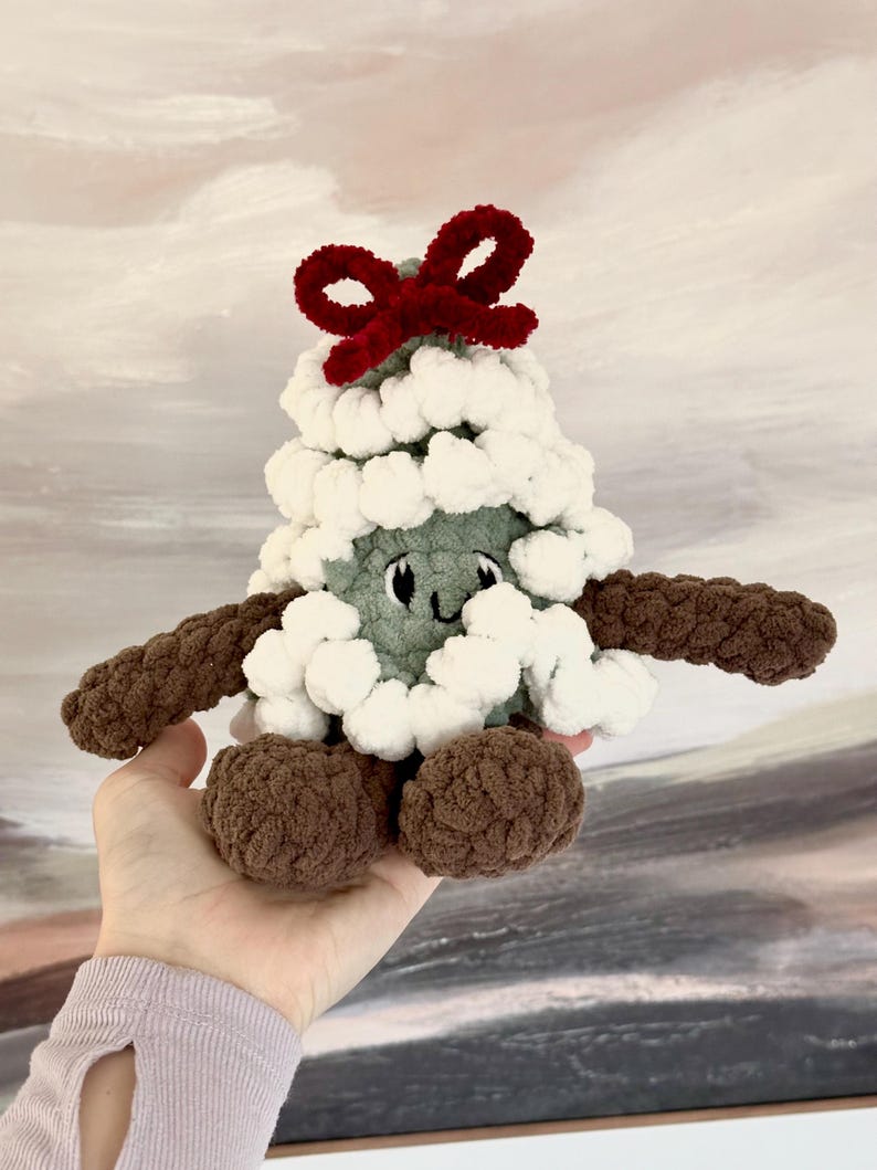 May include: A green crocheted Christmas tree plush toy with white pom-poms and a red bow on top. The toy has a smiling face and brown crocheted arms and legs.