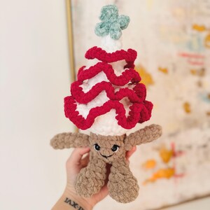 May include: A crocheted plush toy shaped like a person wearing a red and white crocheted Christmas tree hat. The toy has a brown fuzzy body and a smiling face.