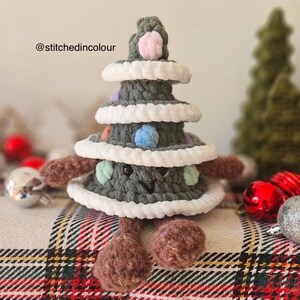 May include: A green crocheted Christmas tree with a smiling face, brown legs, and colorful pom-poms. The tree is sitting on a plaid fabric.