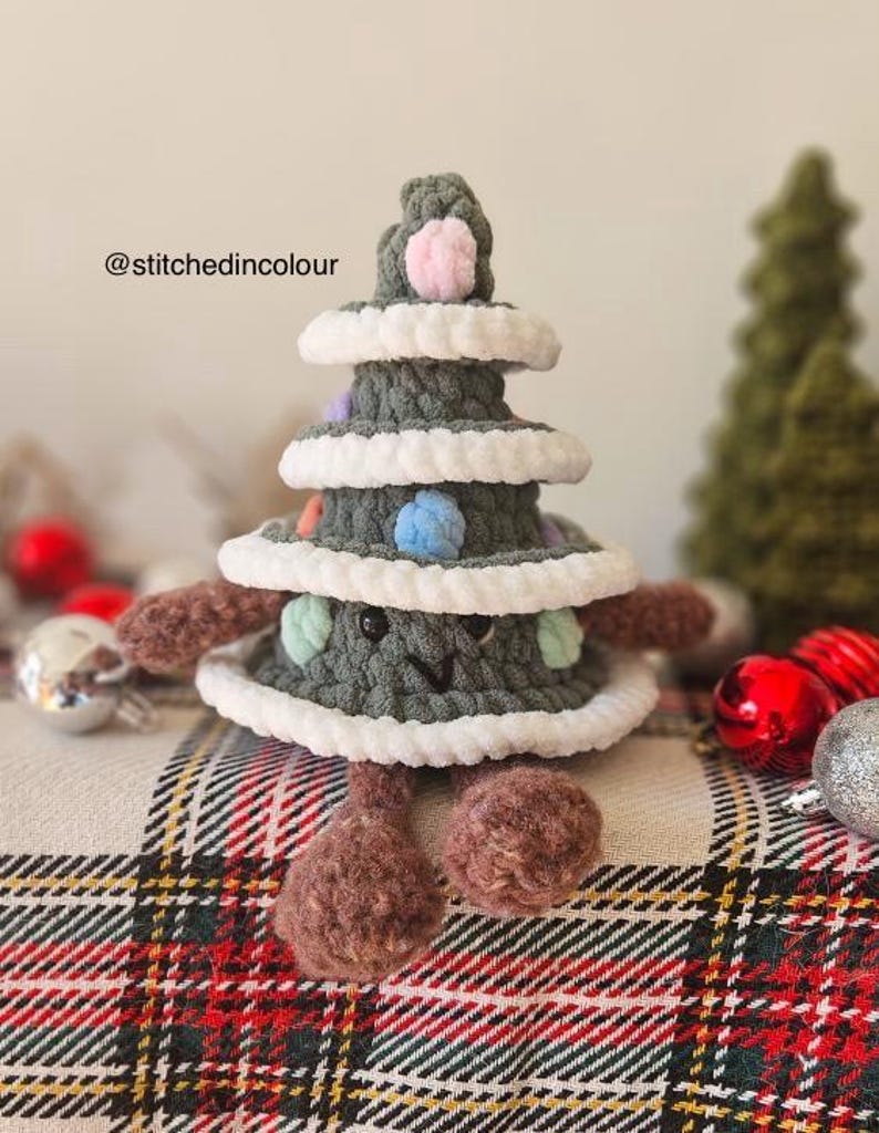 May include: A green crocheted Christmas tree with a smiling face, brown legs, and colorful pom-poms. The tree is sitting on a plaid fabric.
