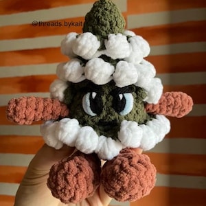 May include: A green crocheted Christmas tree with white crocheted snowballs and a grumpy face. The tree has brown crocheted arms and legs.