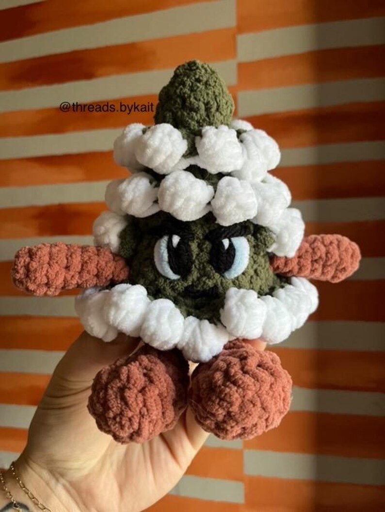 May include: A green crocheted Christmas tree with white crocheted snowballs and a grumpy face. The tree has brown crocheted arms and legs.