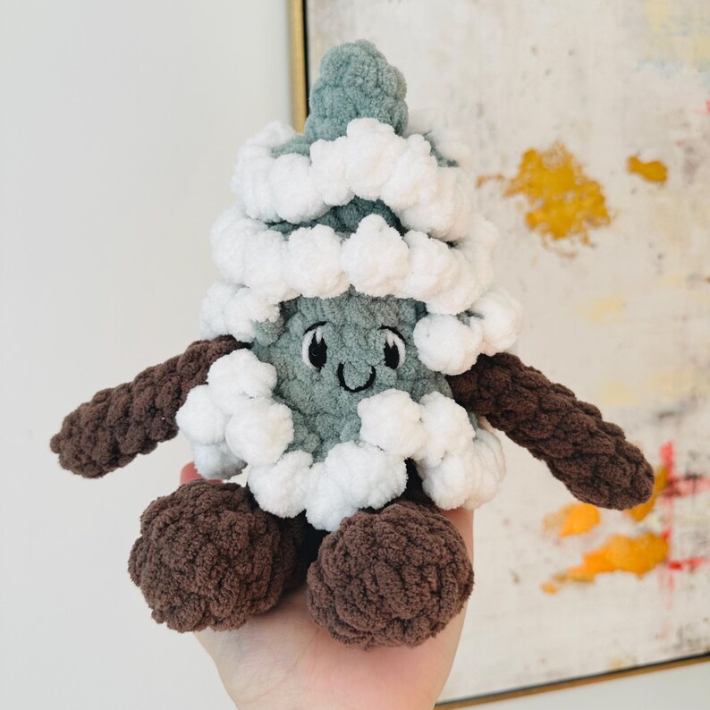 May include: A crocheted plush toy shaped like a Christmas tree with a smiling face. The tree is green with white pom-poms and brown arms and legs.