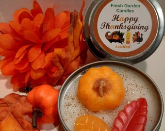 May include: A small tin candle with a white wax base and a pumpkin, leaves, and a red leaf on top. The tin is labeled "Fresh Garden Candles Happy Thanksgiving scented".