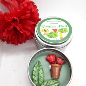 May include: A silver tin candle with a green lid and a label that reads "Fresh Garden Candles Garden Mint Scented". The candle is decorated with a small pot of red flowers and green leaves.