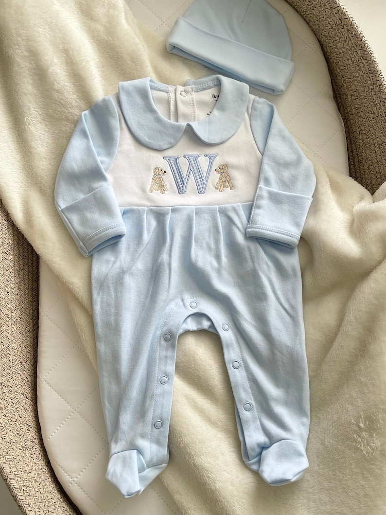 May include: A light blue baby onesie with a Peter Pan collar and matching hat. The onesie features the letter "W" embroidered in blue, flanked by two dog illustrations. The outfit is made of soft fabric and has snap closures.