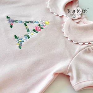 May include: A light pink baby bodysuit with a floral embroidered letter "T". The bodysuit has a Peter Pan collar with a scalloped edge and short sleeves. The text "Tiny Wolfe BABY BOUTIQUE" is printed on the right side.