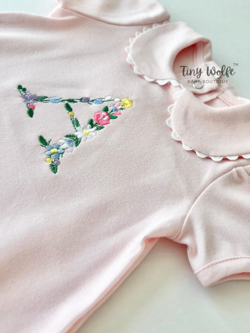 May include: A light pink baby bodysuit with a floral embroidered letter "T". The bodysuit has a Peter Pan collar with a scalloped edge and short sleeves. The text "Tiny Wolfe BABY BOUTIQUE" is printed on the right side.