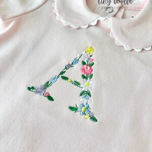 May include: A light pink baby bodysuit with a white scalloped collar. The letter "A" is embroidered with colorful flowers. The text "Tiny Wolfe Baby Boutique" is printed on the top.