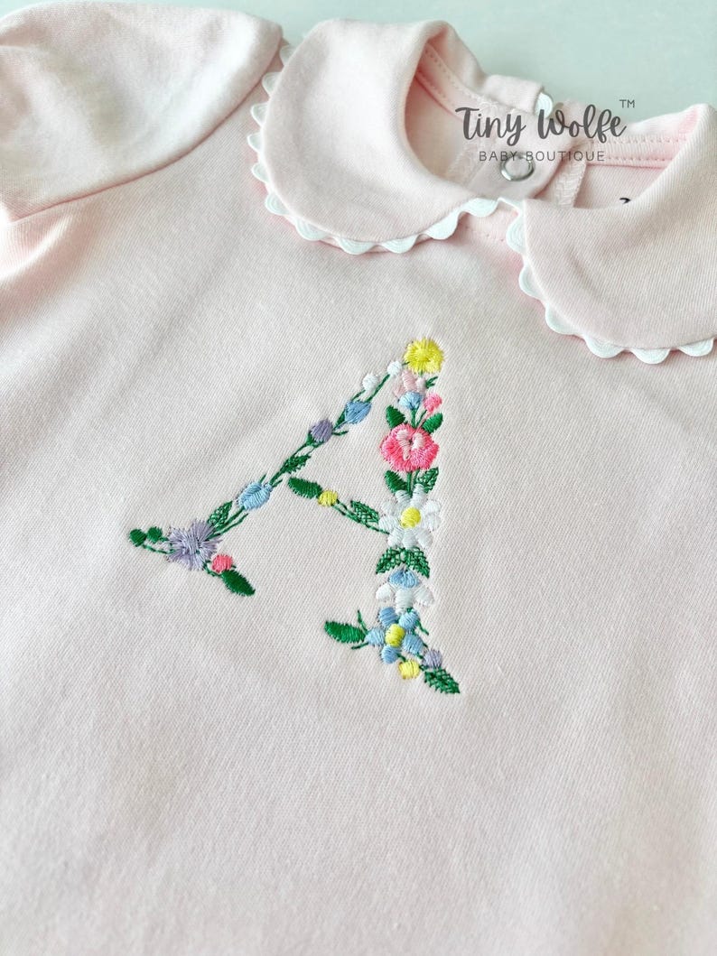 May include: A light pink baby bodysuit with a white scalloped collar. The letter "A" is embroidered with colorful flowers. The text "Tiny Wolfe Baby Boutique" is printed on the top.