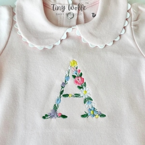 May include: A light pink baby bodysuit with a Peter Pan collar trimmed with white scalloped lace. The letter "A" is embroidered on the front with colorful flowers. The text "Tiny Wolfe Baby Boutique" is printed on the collar.