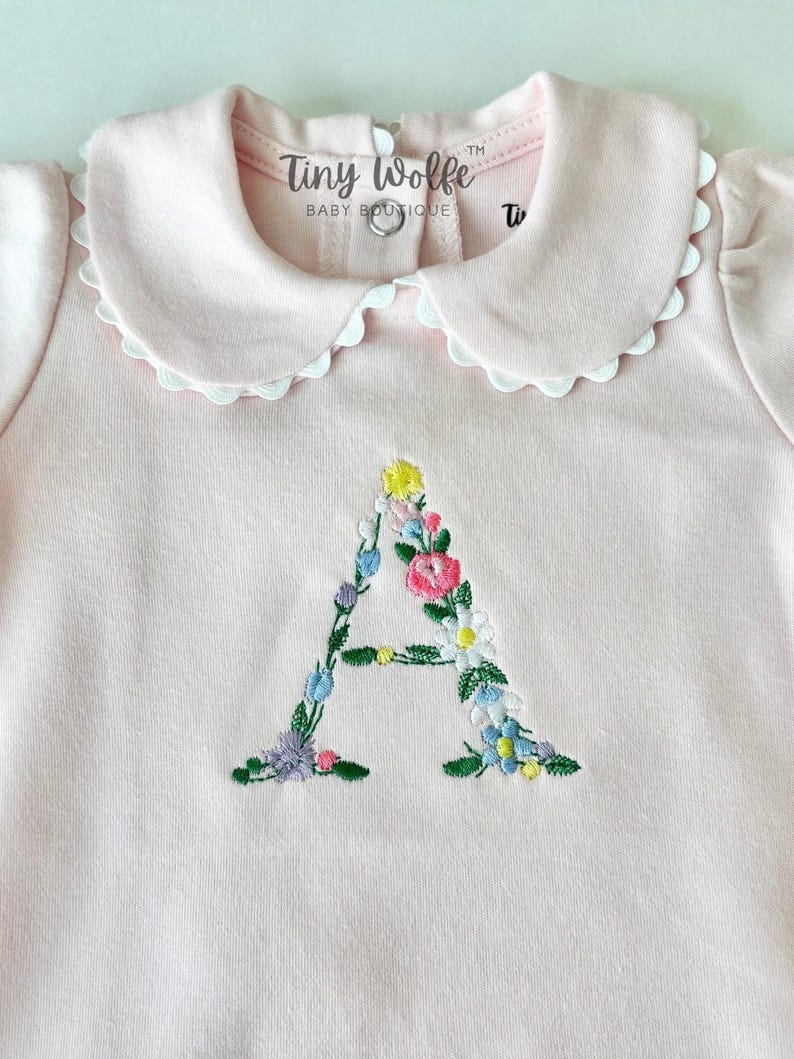 May include: A light pink baby bodysuit with a Peter Pan collar trimmed with white scalloped lace. The letter "A" is embroidered on the front with colorful flowers. The text "Tiny Wolfe Baby Boutique" is printed on the collar.