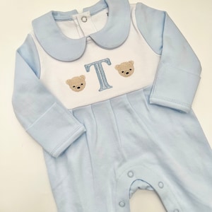 May include: A light blue baby romper with a white bib-style front. The romper features a light blue collar and long sleeves. Embroidered on the front are two teddy bear faces flanking a light blue letter "T". The romper has snap closures down the front.