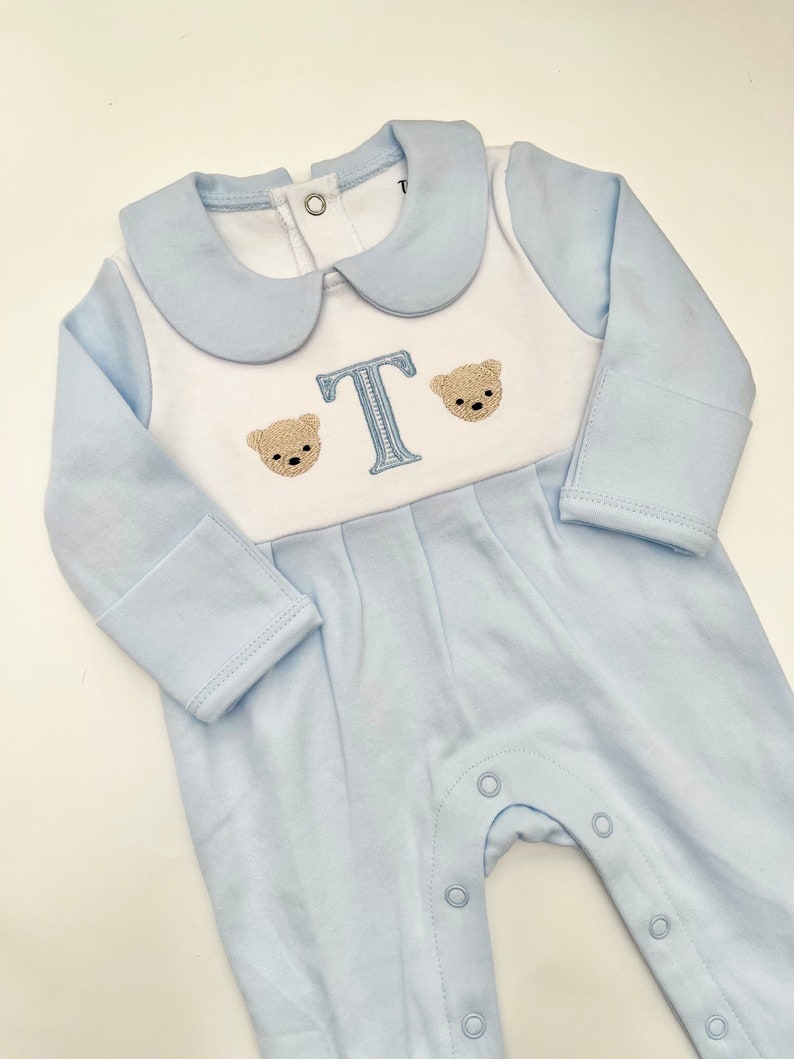 May include: A light blue baby romper with a white bib-style front. The romper features a light blue collar and long sleeves. Embroidered on the front are two teddy bear faces flanking a light blue letter "T". The romper has snap closures down the front.