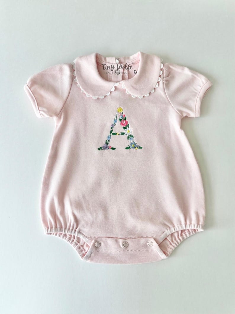 May include: A light pink baby romper with a white scalloped collar and short sleeves. The letter "A" is embroidered on the front with colorful flowers. The brand name "Tiny Wolfe" is printed on the collar.