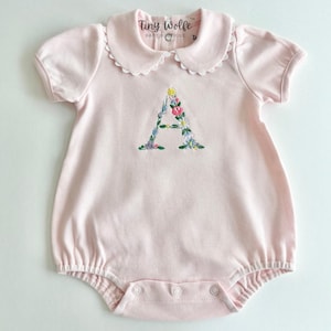May include: A light pink baby romper with a scalloped collar and short sleeves. The romper features an embroidered floral "A" on the front. The brand name "Tiny Wolfe Baby Boutique" is printed on the collar.