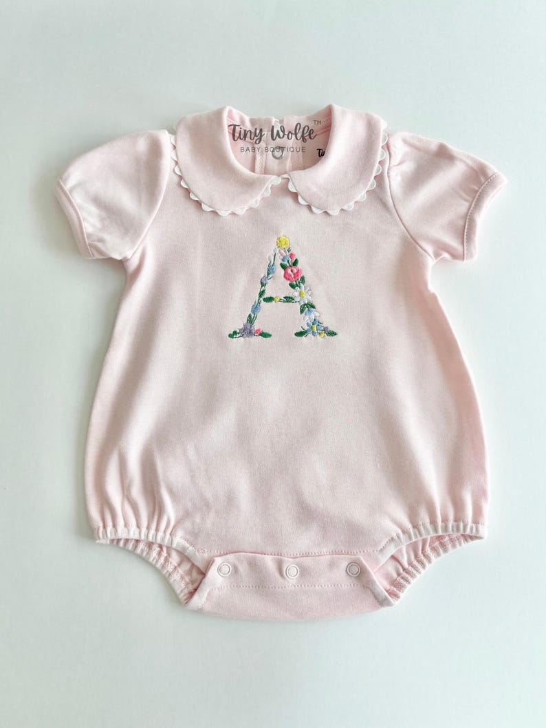 May include: A light pink baby romper with a scalloped collar and short sleeves. The romper features an embroidered floral "A" on the front. The brand name "Tiny Wolfe Baby Boutique" is printed on the collar.