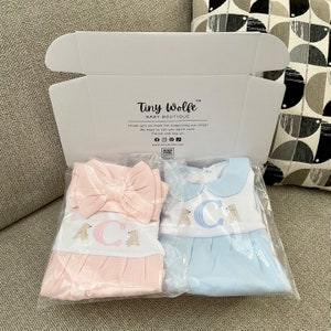May include: A white box from Tiny Wolfe Baby Boutique contains two baby outfits. One is pink with a matching bow, the other is blue. Both outfits have a white top with a letter "C" and dog embroidery.