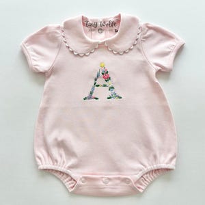 May include: A light pink baby romper with a Peter Pan collar and short sleeves. The letter "A" is embroidered on the front with colorful flowers. The tag reads "Tiny Wolfe Baby Boutique".