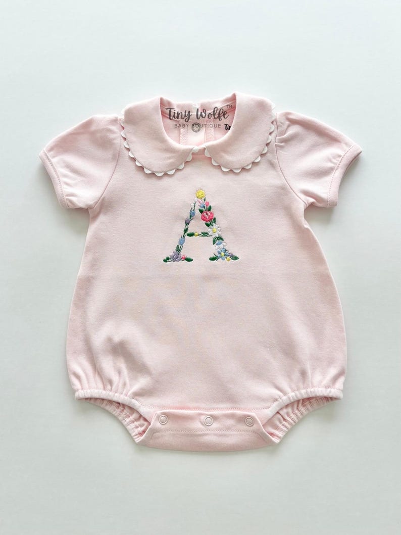 May include: A light pink baby romper with a Peter Pan collar and short sleeves. The letter "A" is embroidered on the front with colorful flowers. The tag reads "Tiny Wolfe Baby Boutique".