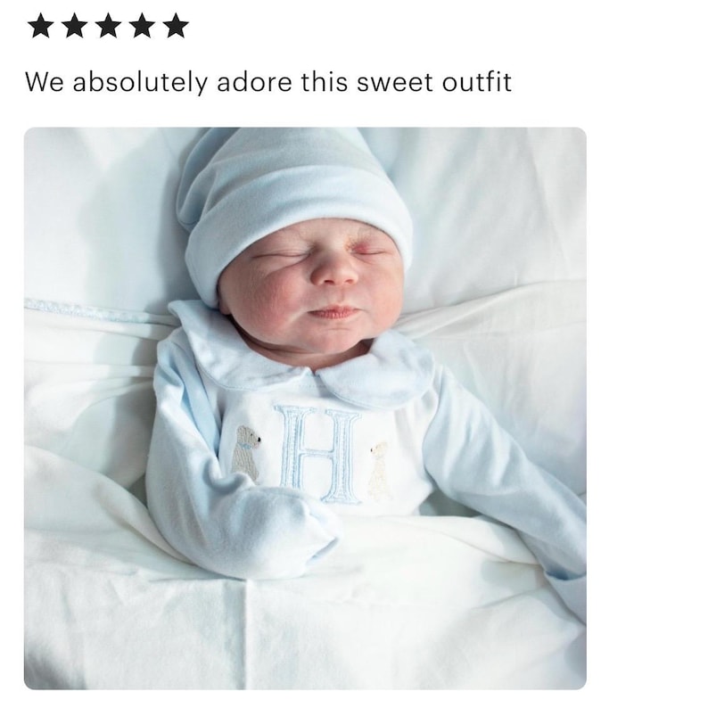 May include: A newborn baby wearing a light blue outfit and matching cap. The outfit features a Peter Pan collar, long sleeves, and an embroidered "H" with two animal figures. The text "We absolutely adore this sweet outfit" is above the baby.